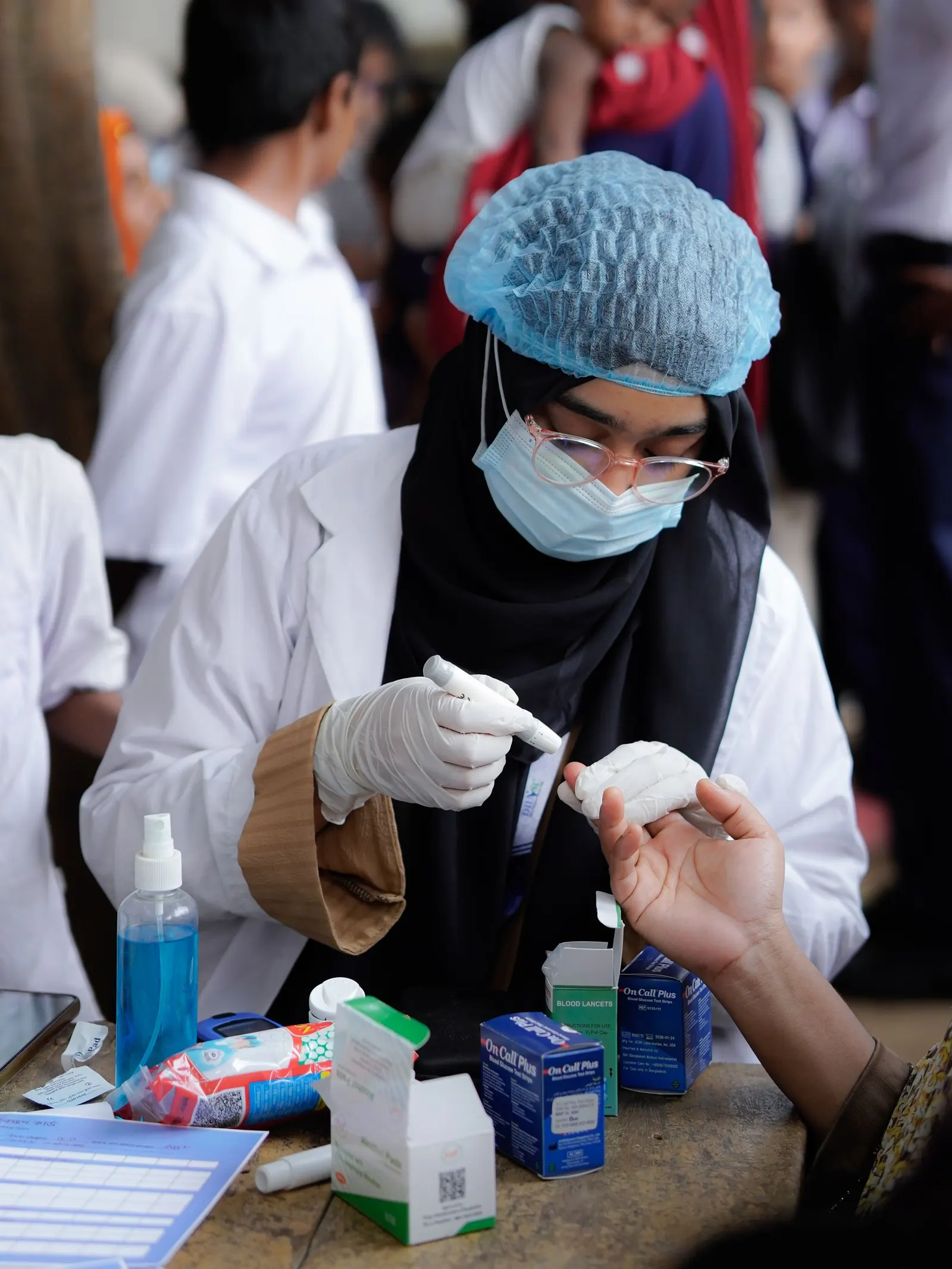 Health worker in a lab coat and hair cover preparing a finger-prick blood test at a screening desk